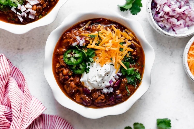 Close up overhead view beef and bean chili in white bowl topped with sour cream, shredded cheddar cheese and jalapeño slices.