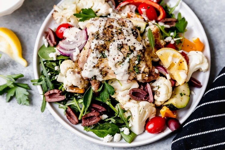 Close up view sheet pan Mediterranean chicken and veggies on white plate with lemon tahini dressing drizzled over top.
