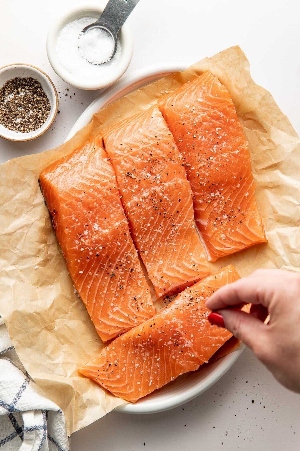 Overhead view of a hand sprinkling salt and pepper over raw salmon fillets on a parchment lined plate.