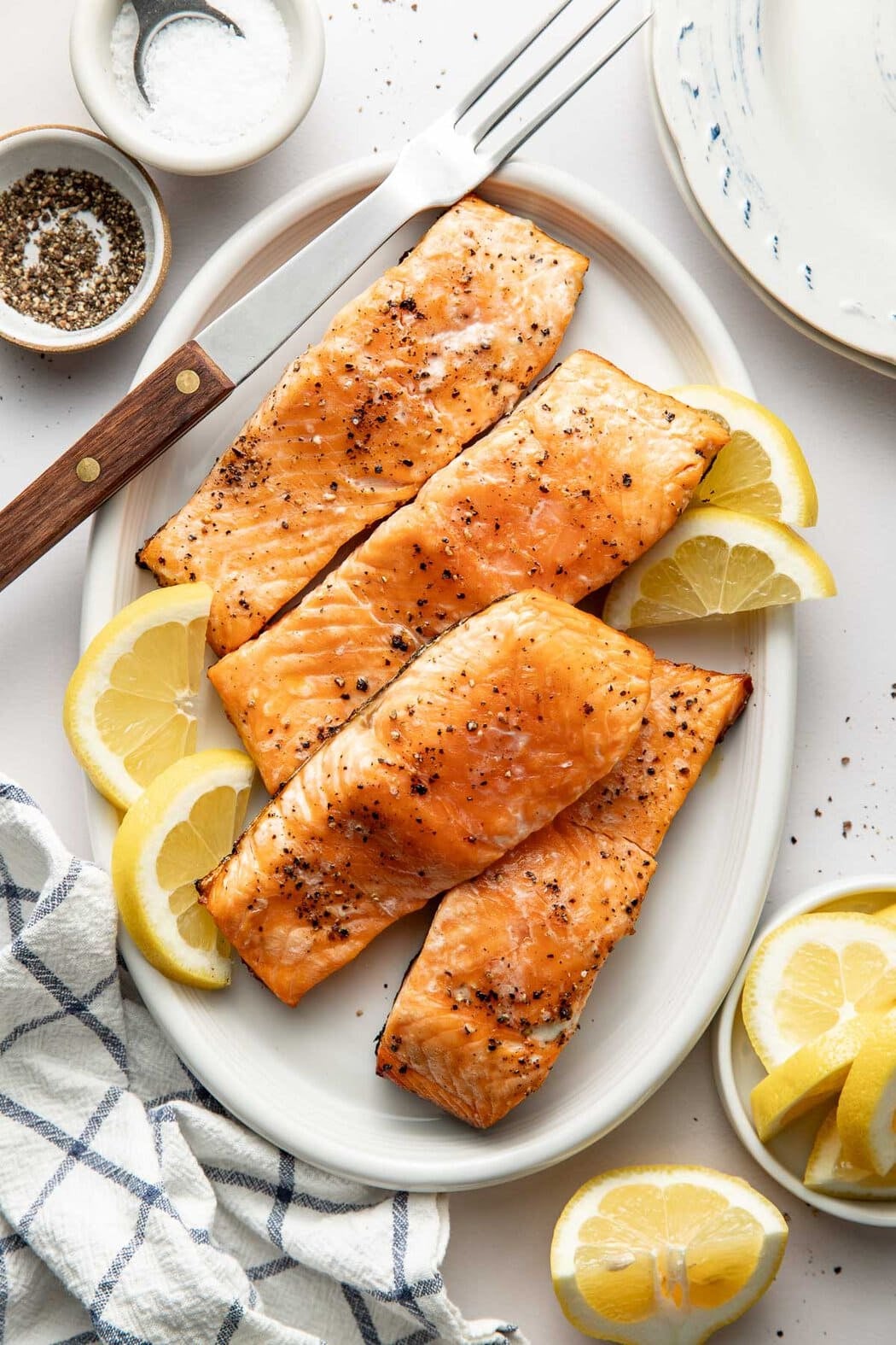Overhead view of an oval shaped platter filled with Grilled Salmon fillets garnished with lemons and sprinkled with black pepper.
