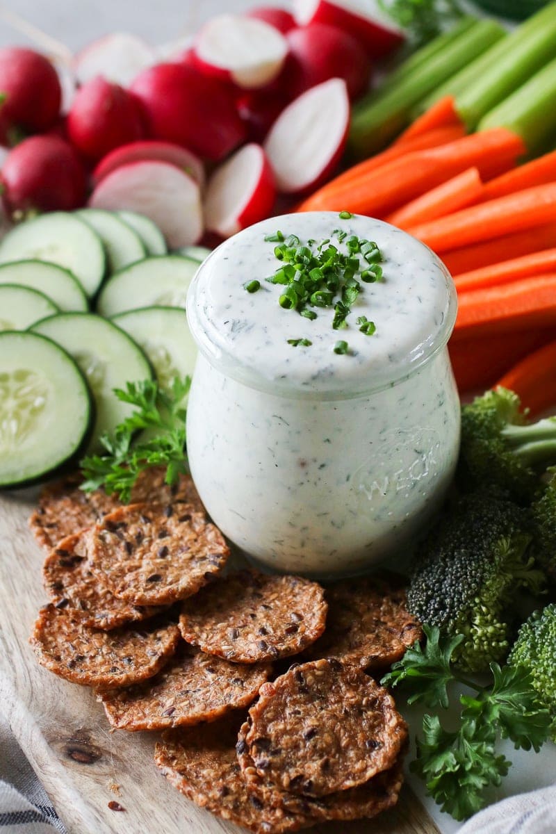 Photo of Healthy Homemade Ranch Dressing in a mason jar topped with fresh chives and surrounded by fresh cut vegetables.