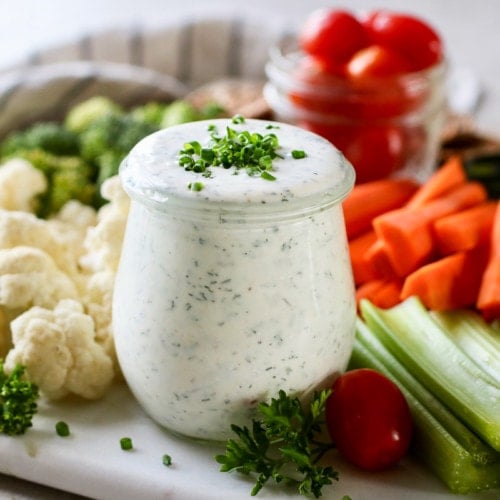 Small jar of homemade ranch dressing on cutting board surrounded by fresh cut vegetables