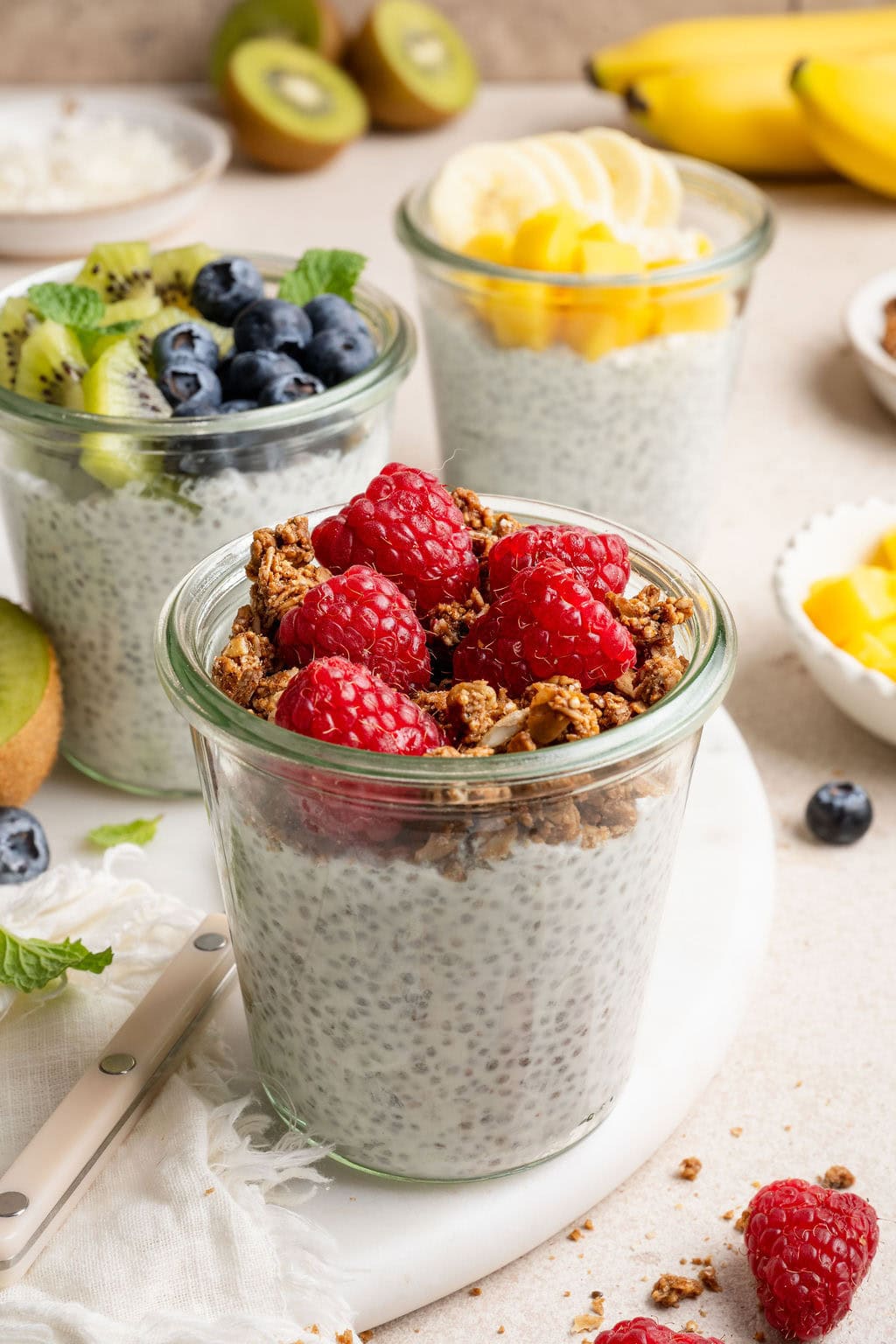 A variety of glass jars filled with chia pudding and topped with colorful fresh fruit and granola on a white marble countertop.