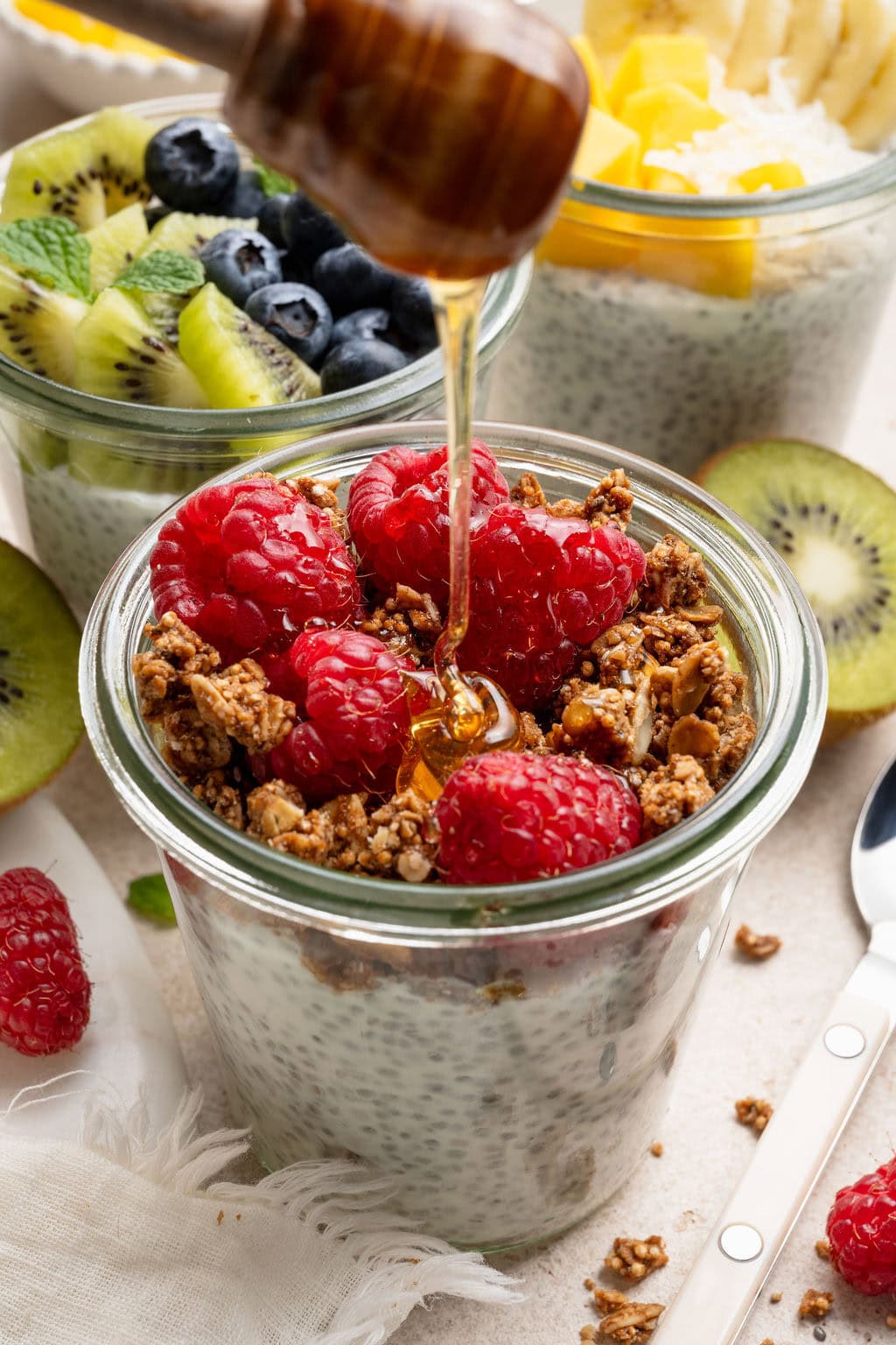 Close up view of honey being poured over a glass jar of chia pudding topped with fresh raspberries and granola. 