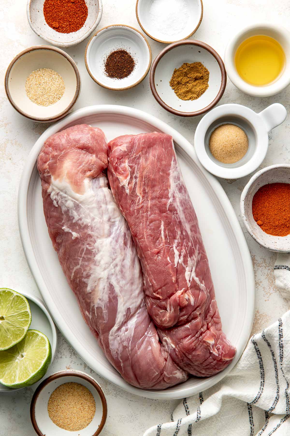 Overhead shot of 2 uncooked pork tenderloins surrounded by small white bowls filled with various spices and 2 lime halves.