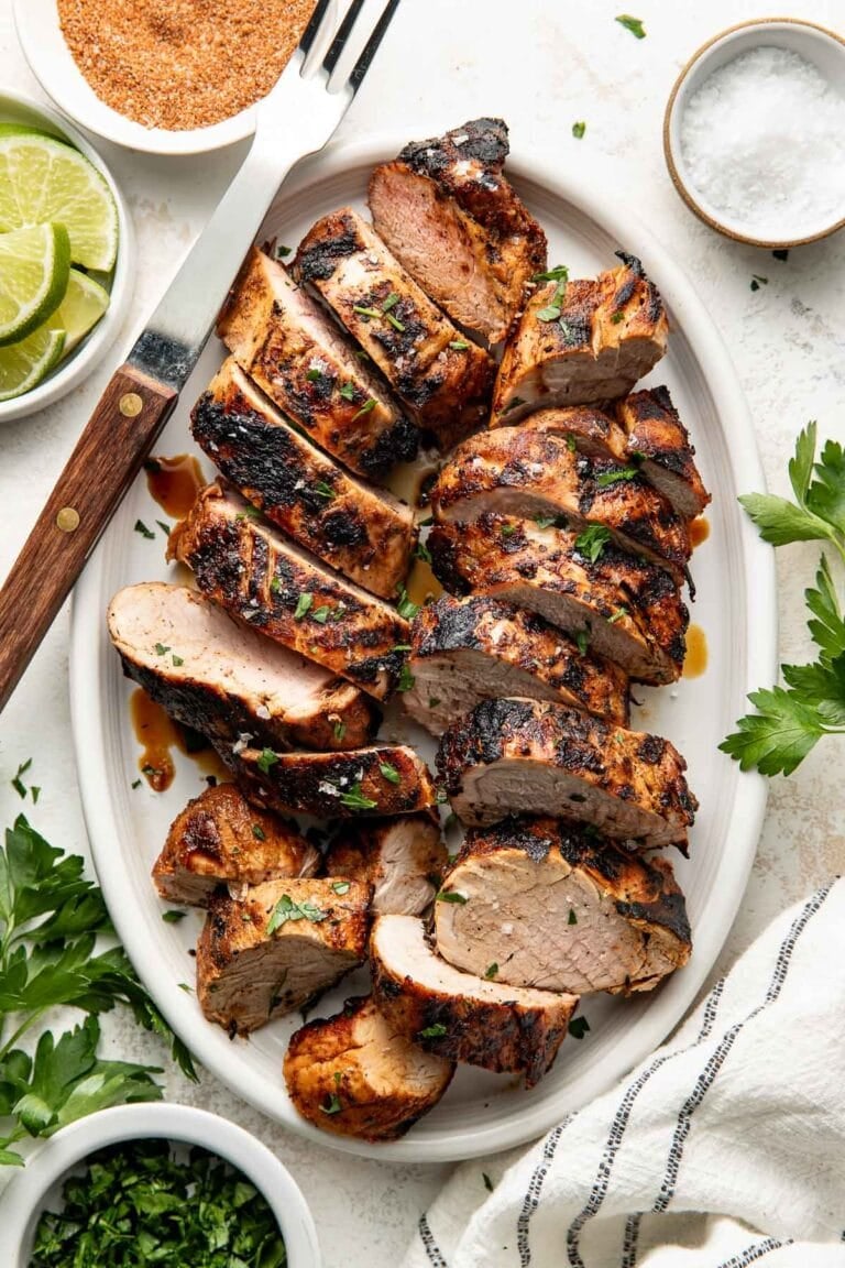 Overhead view of sliced pork tenderloin that has been perfectly grilled and arranged on a large white oval platter with a serving fork on the side.