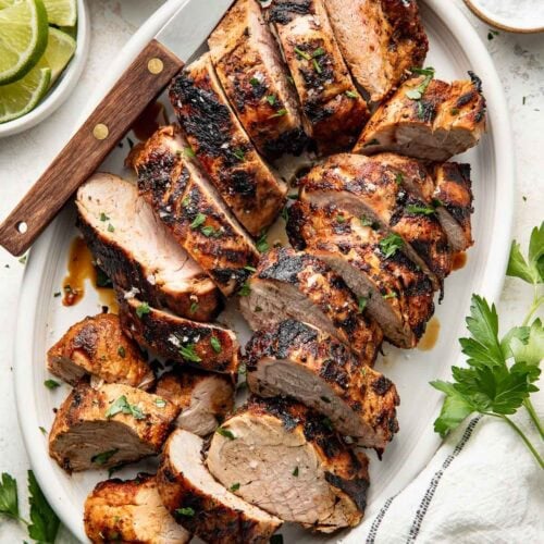 Perfectly grilled and sliced pork tenderloins arranged on a white oval platter surrounded by fresh parsley, limes and red spice mixture in a white bowl.
