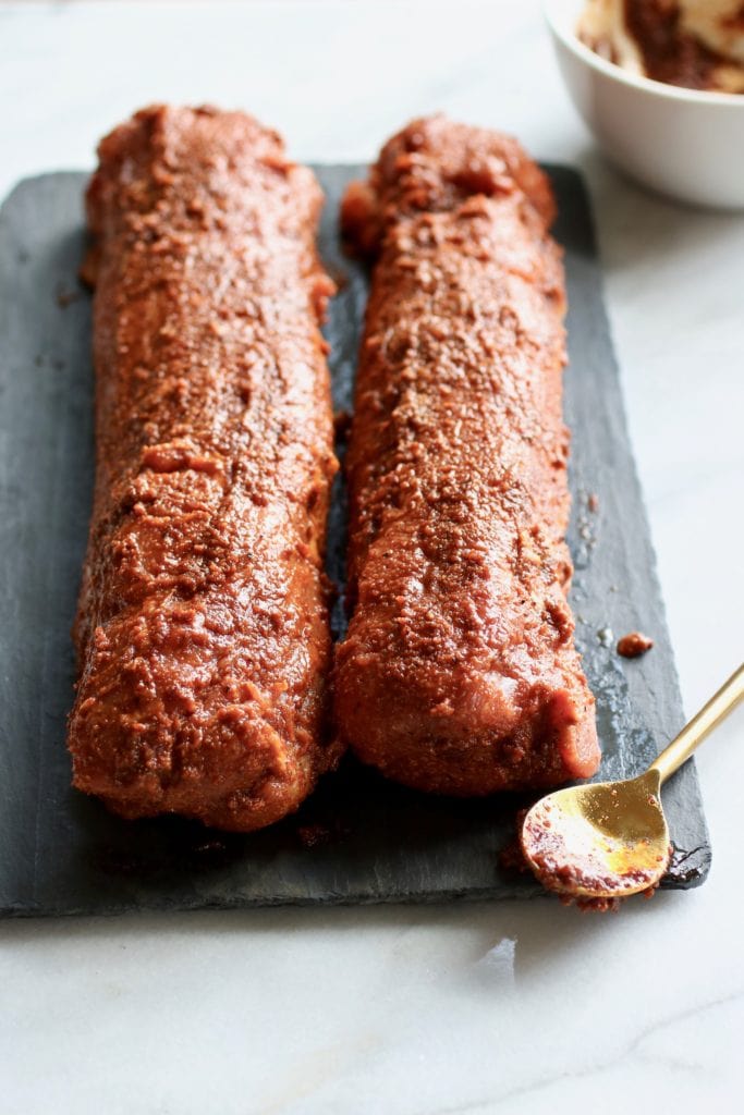 Two pork tenderloin on a black serving board smeared with the Ultimate BBQ Rub.