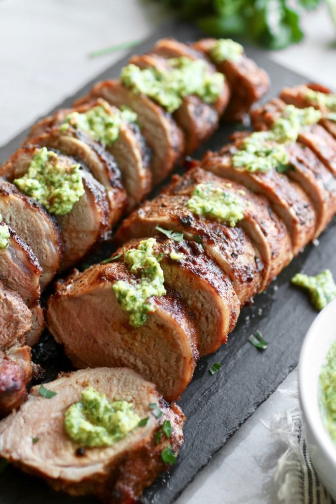 Photo of Grilled Pork Tenderloin on a black serving board topped with Avocado Green Sauce.