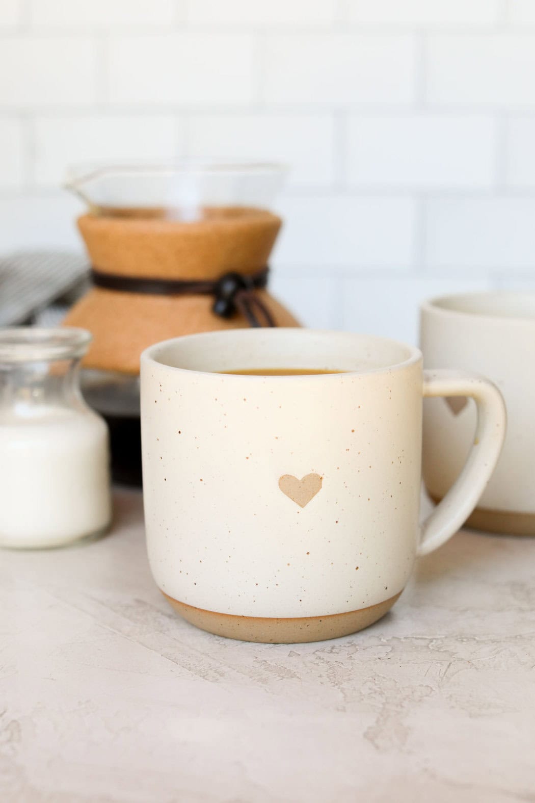 White coffee mug with small heart on it filled with coffee and cream in the background. 