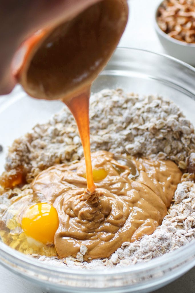A hand pours peanut butter into a bowl with oats, flour, brown sugar and eggs.