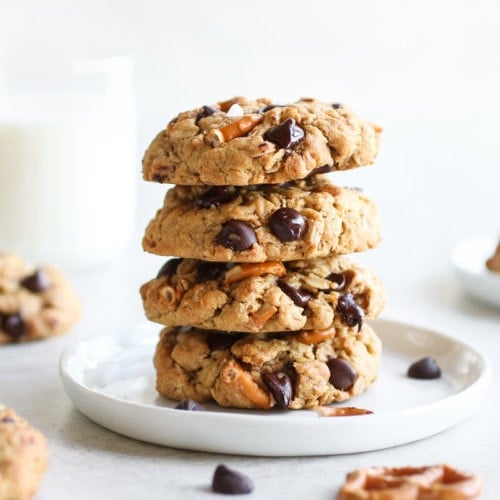A stack of four Peanut Butter Chocolate Chip Pretzel Cookies on a white plate with a glass on milk in the background.