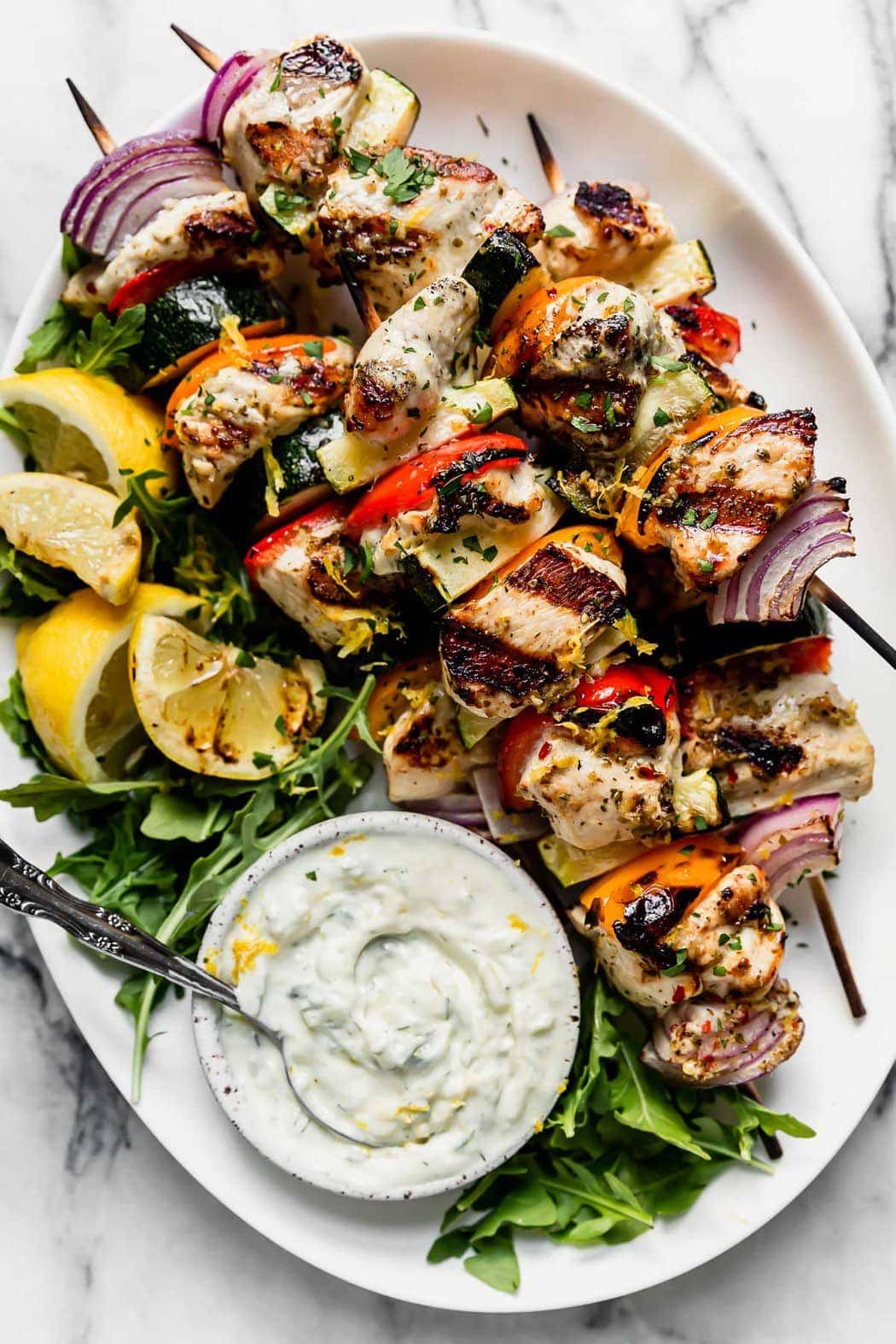 Photo of Greek Chicken Kebabs with Tzatziki Sauce on a serve platter with arugula, grilled lemons and a bowl to Tzatziki sauce.