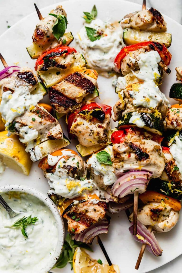 Overhead view of Greek chicken kebabs drizzled with tzatziki sauce and topped with fresh herbs and black peppers. 