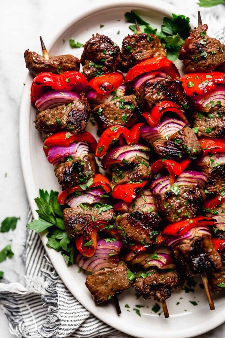Overhead view of an oval platter filled with grilled steak kebabs on skewers showing the layers of steak, red pepper, and red onion.