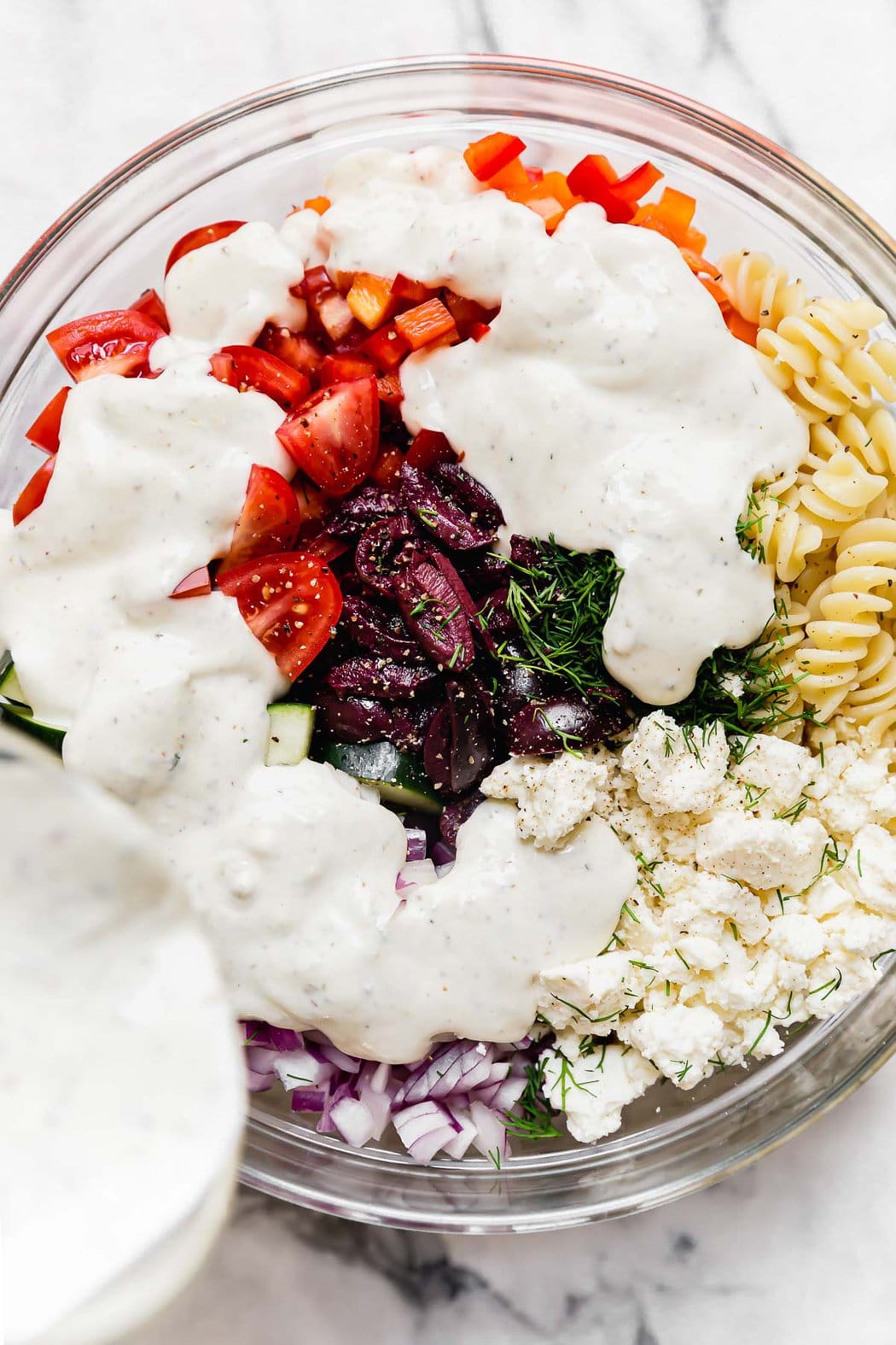 Greek Veggie Pasta Salad ingredients in a bowl with dressing being poured over the top