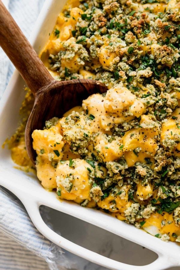 Close view of creamy vegan cauliflower mac and cheese in a white baking dish with a wooden serving spoon scooping up a portion.