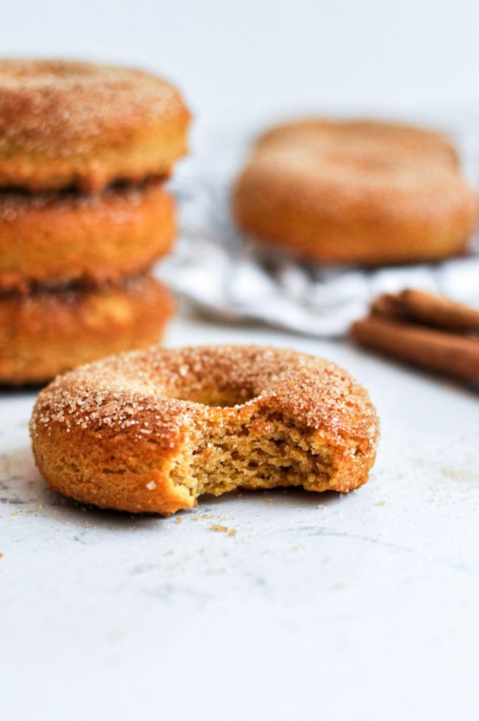 Healthy Baked Pumpkin Donuts with a bit taken out of it with a stack of three pumpkin donuts behind it.