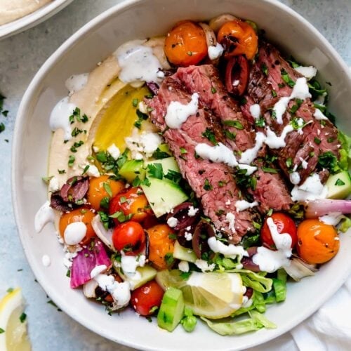 Overhead view of a bowl of hummus, grilled and fresh veggies, and tender slices of flank steak all sprinkled with feta cheese.