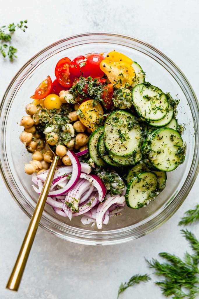 Garbanzo bean salad ingredients for the Mediterranean Bowls in a clear glass bowl. Includes sliced red onion, cherry tomatoes, sliced cucumber, garbanzo beans, herbs and oil.