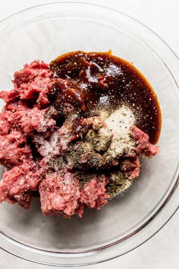 Raw ground beef, seasonings, and bbq sauce in a clear bowl ready to be mixed together