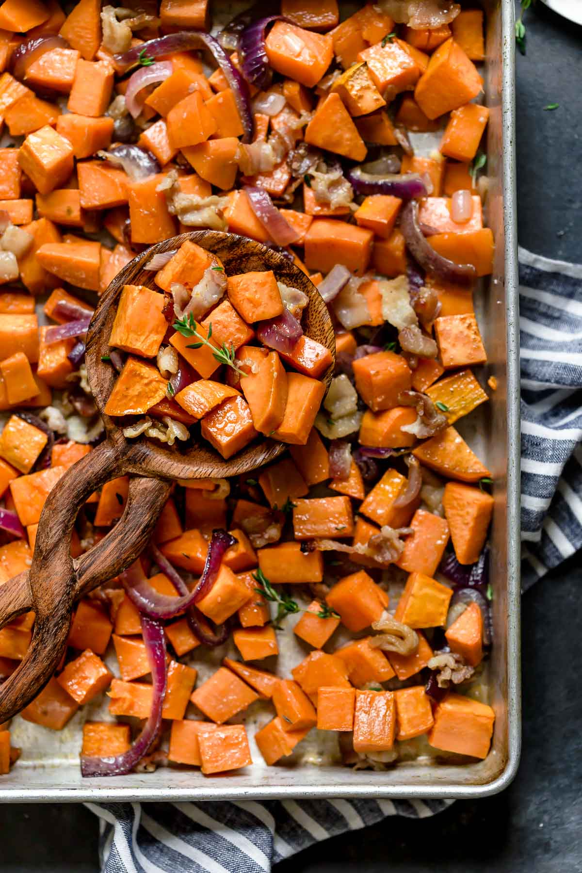 Overhead view of a wooden spoonful of Roasted Sweet Potatoes with Maple and Bacon resting on a sheet pan of them.
