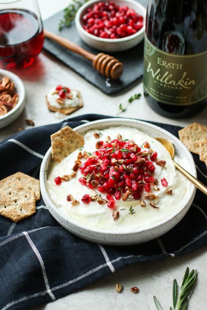 Whipped Goat Cheese in a white serving bowl, topped with pomegranate, fresh thyme and served with crackers.