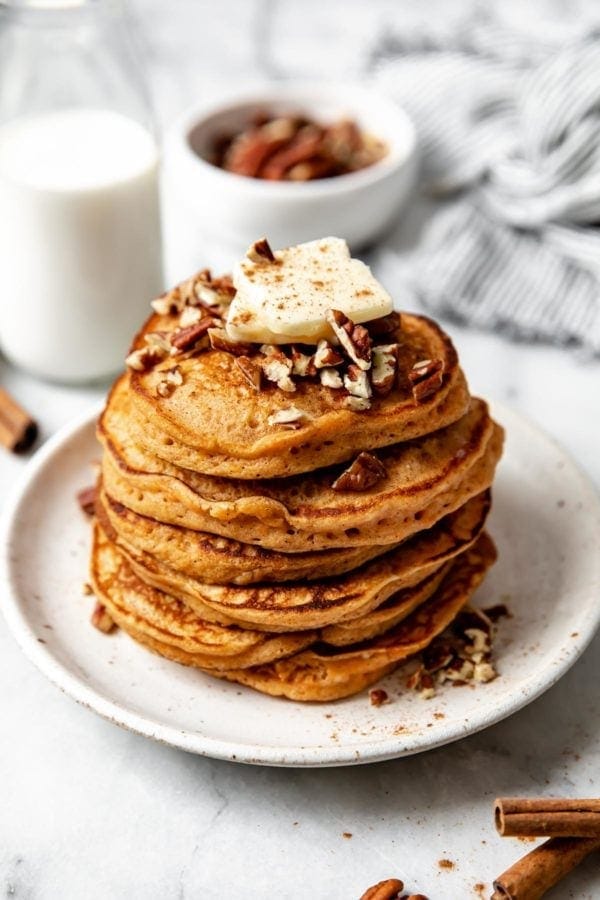 A large stack of fluffy gluten free sweet potato pancakes on a white plate topped with a pad of butter and chopped pecans