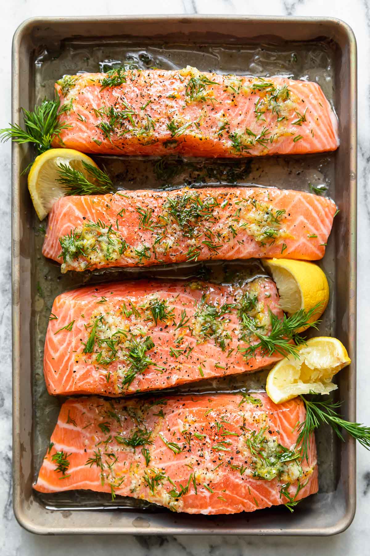 Four raw salmon filets lined up on a sheet pan drizzled with olive oil and fresh herbs.