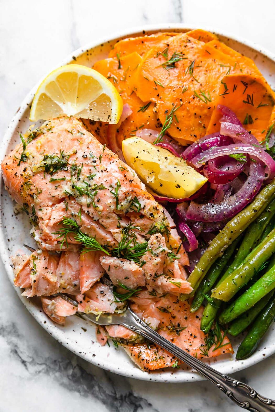 A salmon fillet, green beans, sweet potato slices, and red onion slices, plated on a speckled plate and topped with lemon wedges.