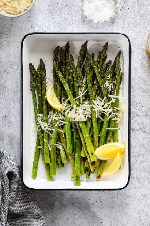 Freshly grilled asparagus spears in a grill pan topped with parmesan cheese and lemon wedges