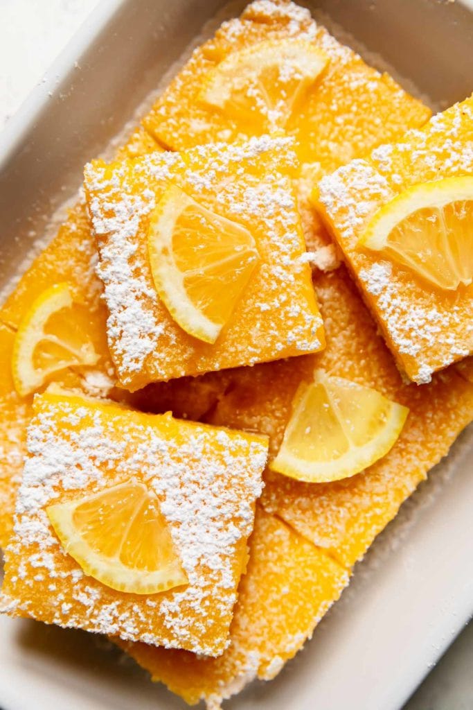 A tray of gluten-free lemon bars sprinkled with powdered sugar and a small lemon slice. 