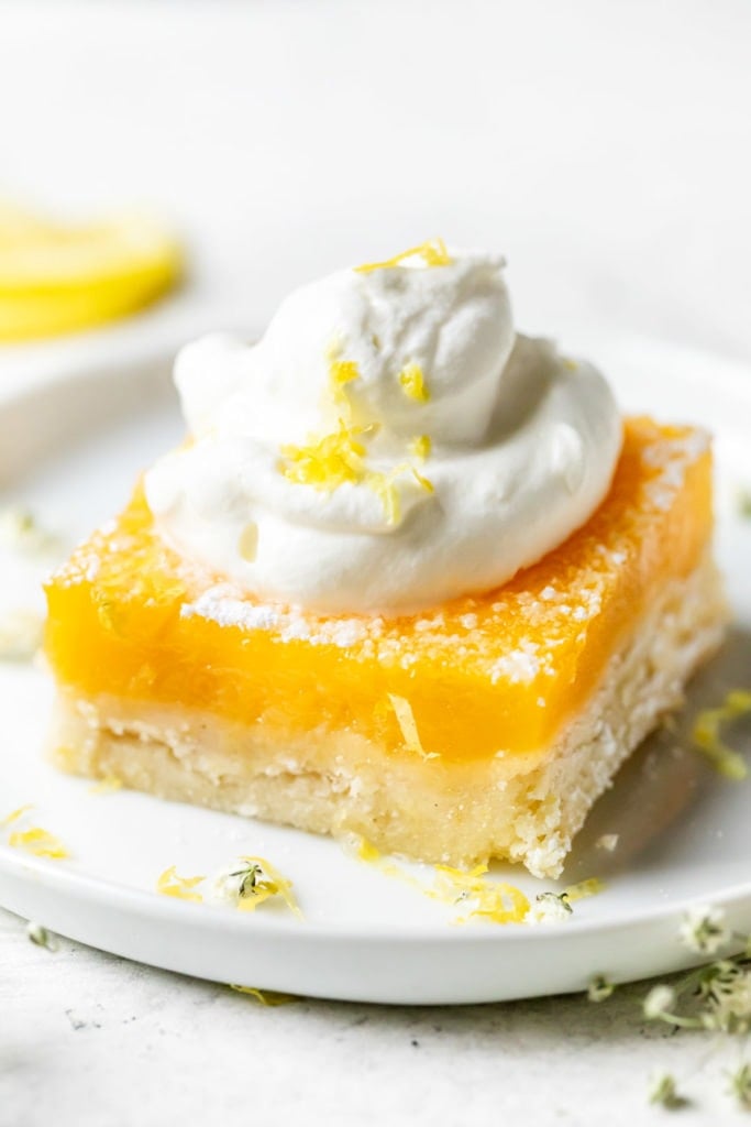 A single lemon bar with a gluten-free crust plated on a small round plate topped high with whipped cream and lemon zest