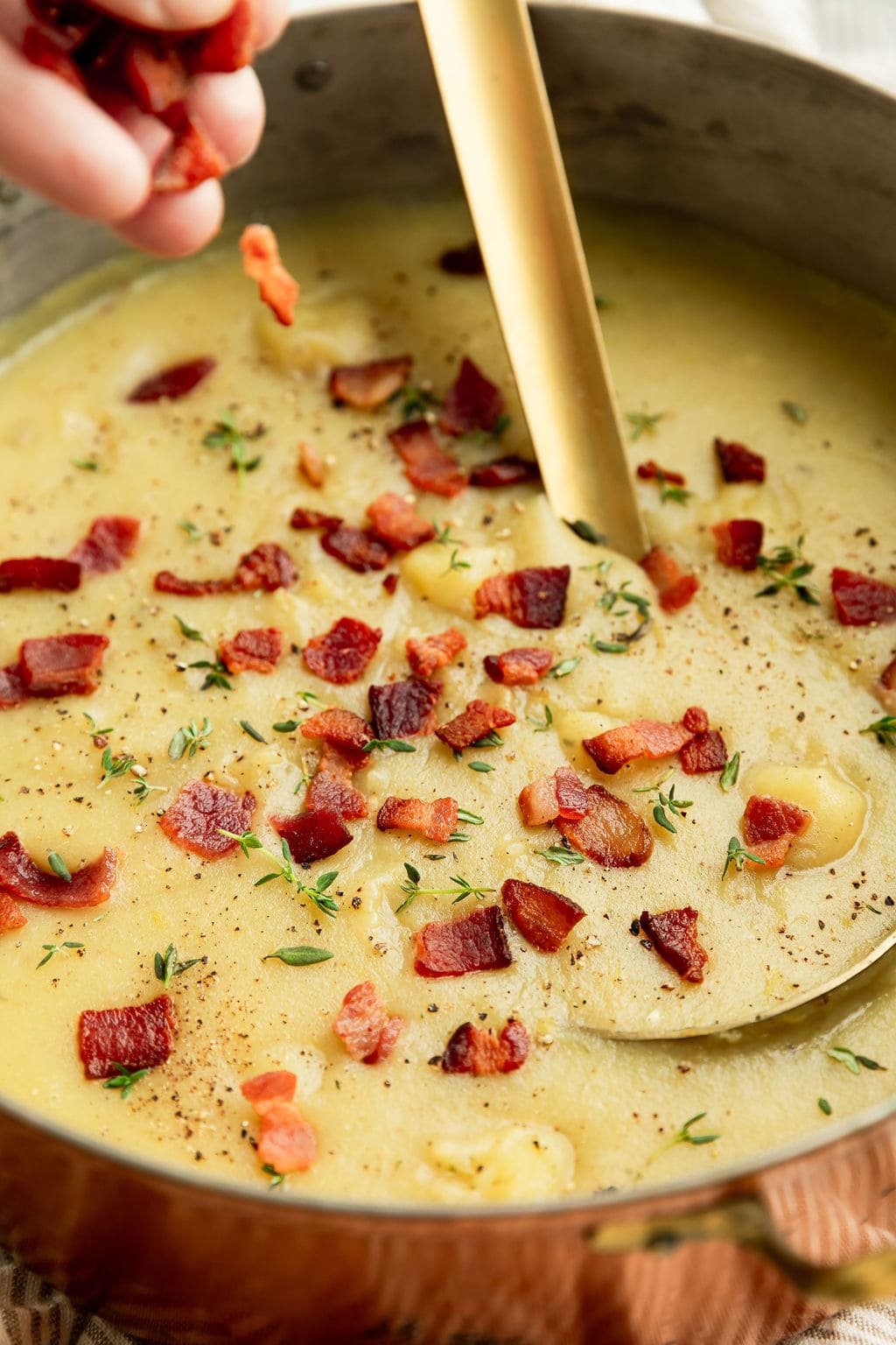 A hand sprinkling cooked bacon onto creamy potato leek soup in a copper soup pot. 