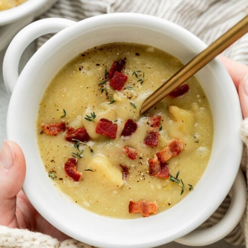 Two hands cradling a small white bowl of creamy potato soup with leeks and bacon.