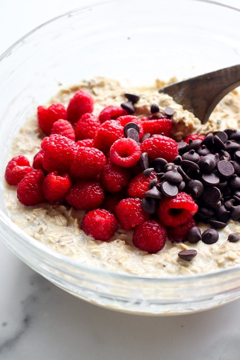 A bowl of oatmeal topped with raspberries and chocolate chips, a wooden spoon sticking out.