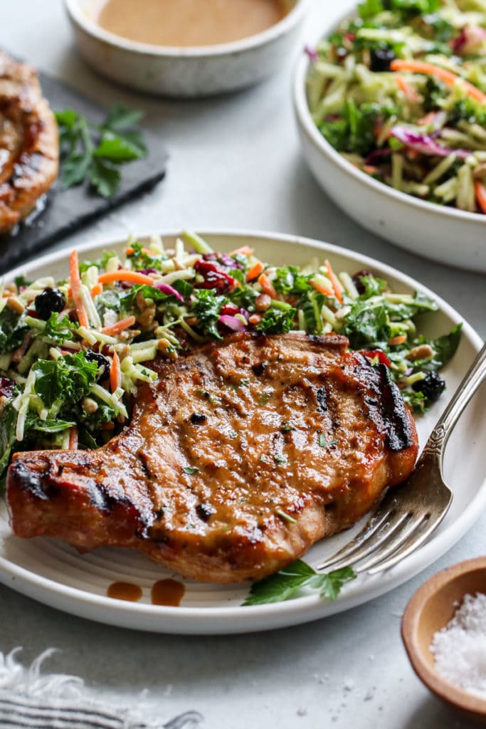 A marinaded grilled bone-in pork chop with grill marks on a white plate with a coleslaw salad on the side. 