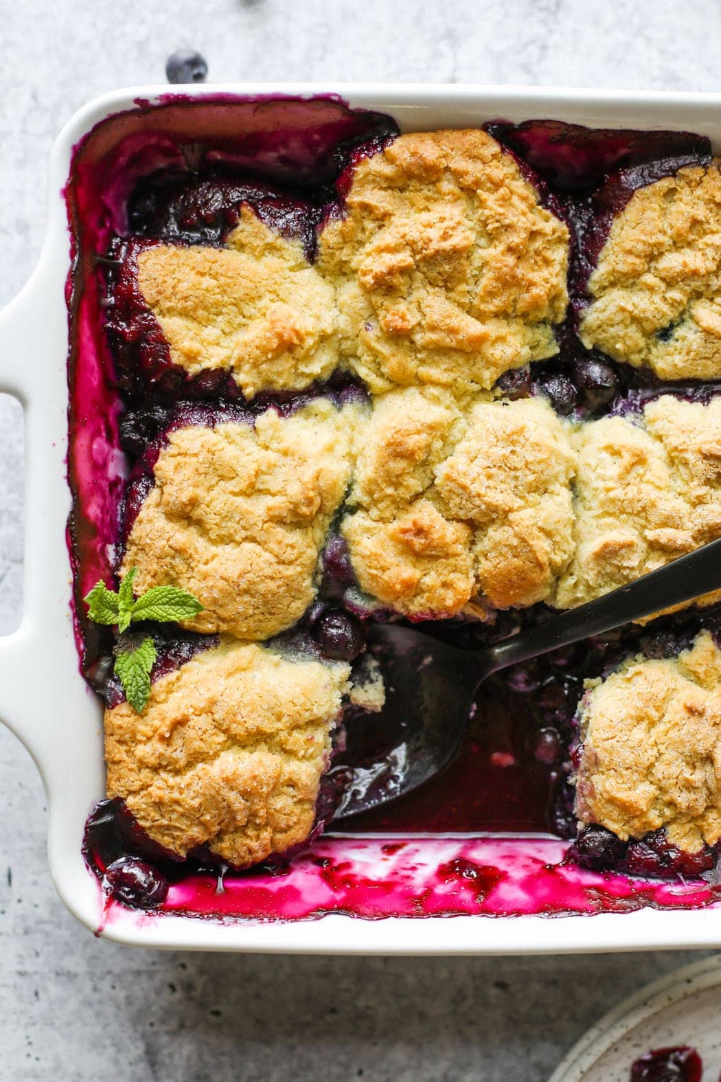 Overhead view white baking dish filled with blueberry cobbler, a black serving spoon in cobbler