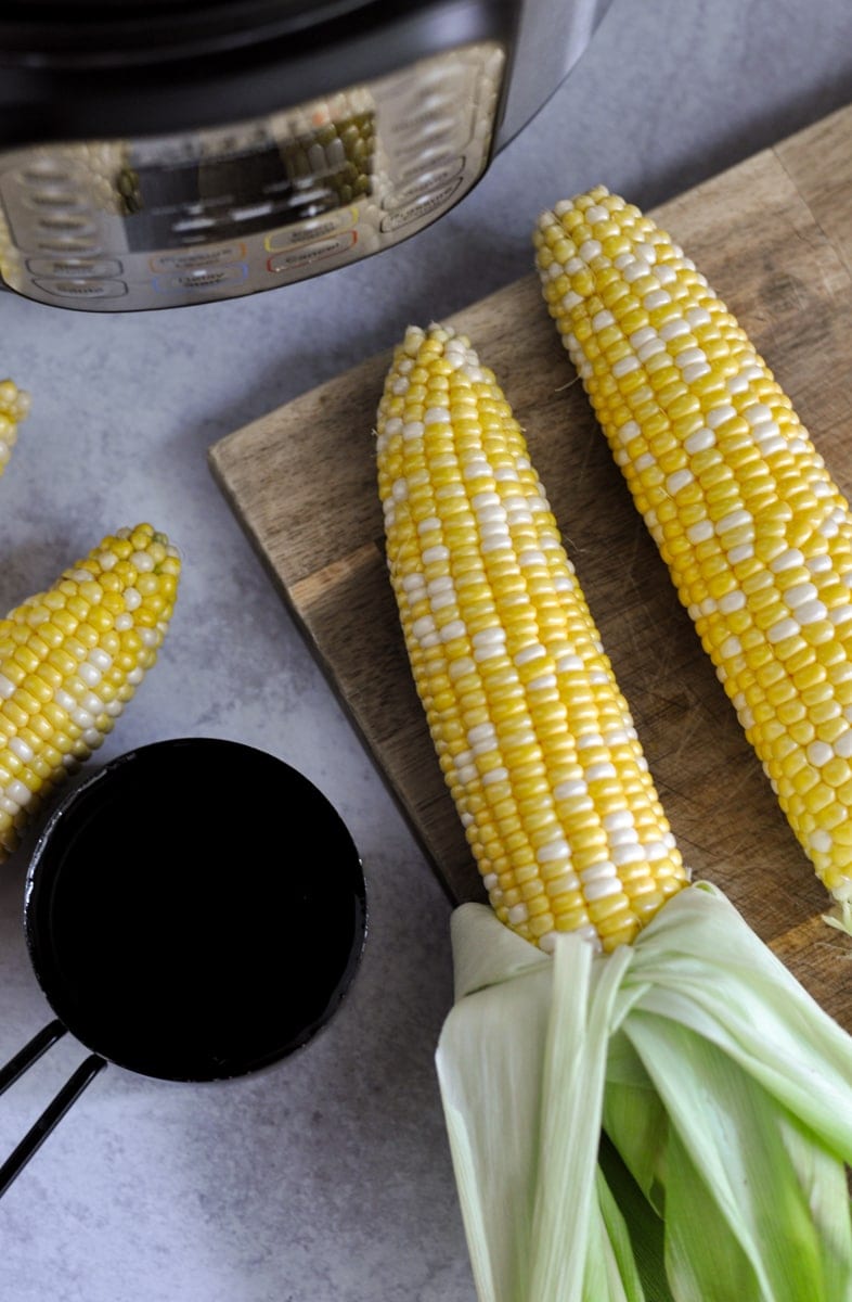 Overhead view of corn on the cob with the green husk pulled down to the end laying on a cutting board next to an Instant Pot and black measuring cup filled with water