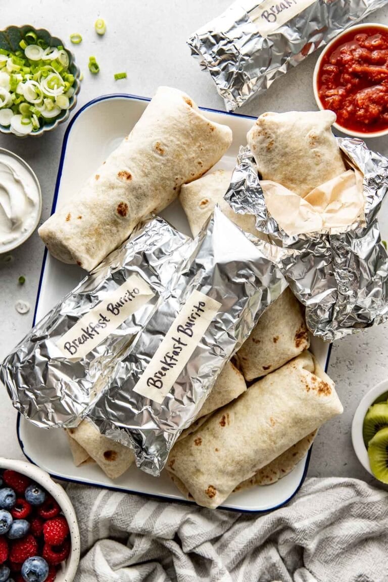Several foil wrapped breakfast burritos on a tray surrounded by toppings including green onions.