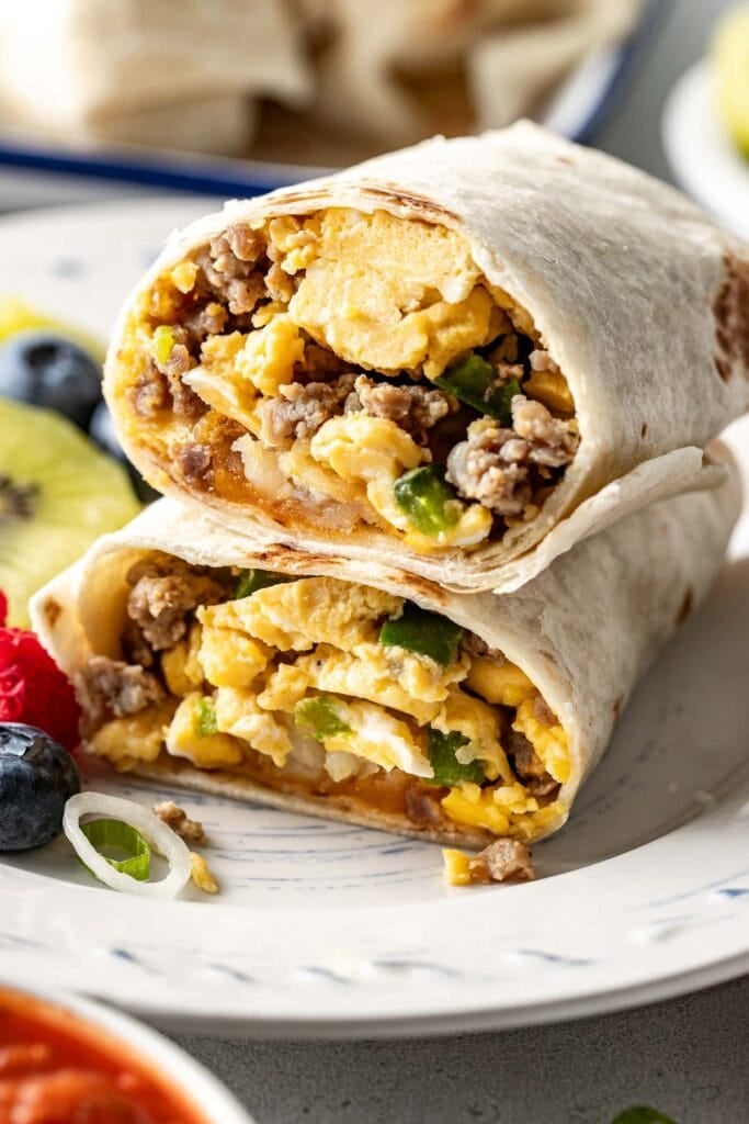 Close up view of a plate filled with a breakfast burrito cut in half showing the egg and sausage mixture on the inside with fruit on the side.