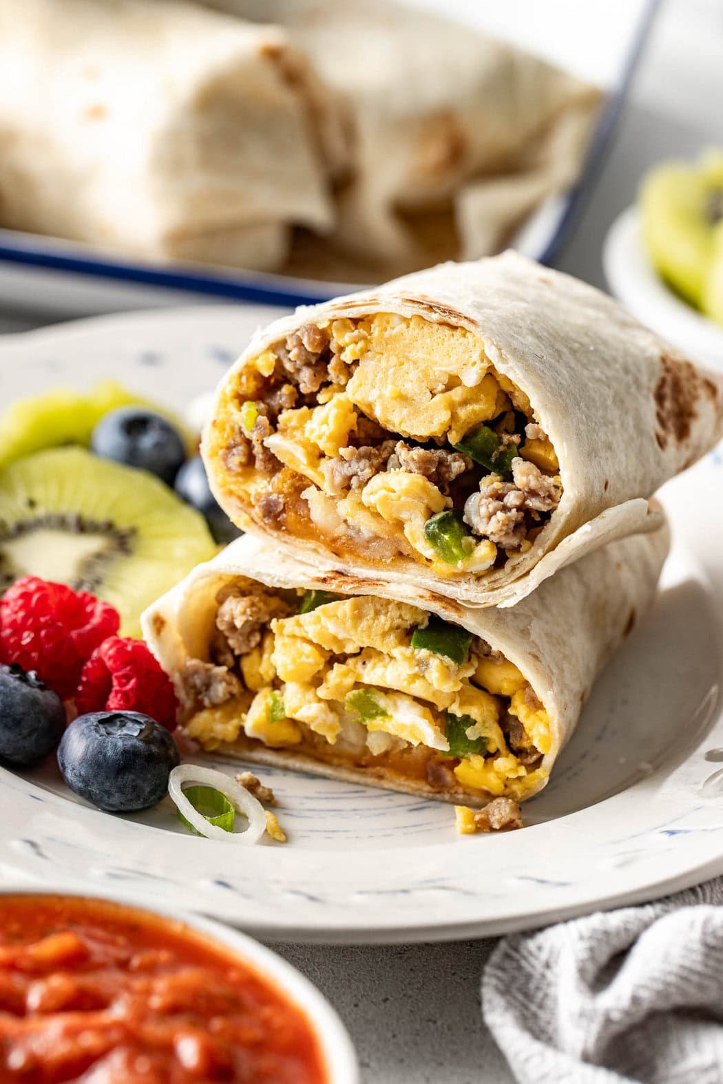 A plate of fresh berries and kiwi that has a breakfast burrito filled with scrambled eggs, peppers, and sausage that has been cut into half.