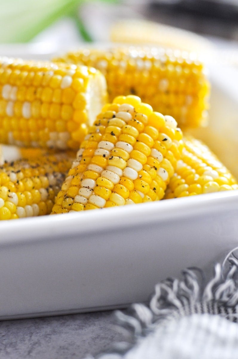 Golden yellow corn on the cob halves in a white serving dish sprinkled with cracked black pepper