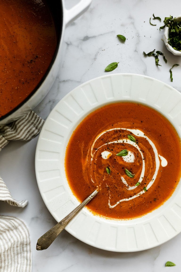 Overhead view of a bowl of creamy tomato soup topped with black pepper and fresh herbs. 