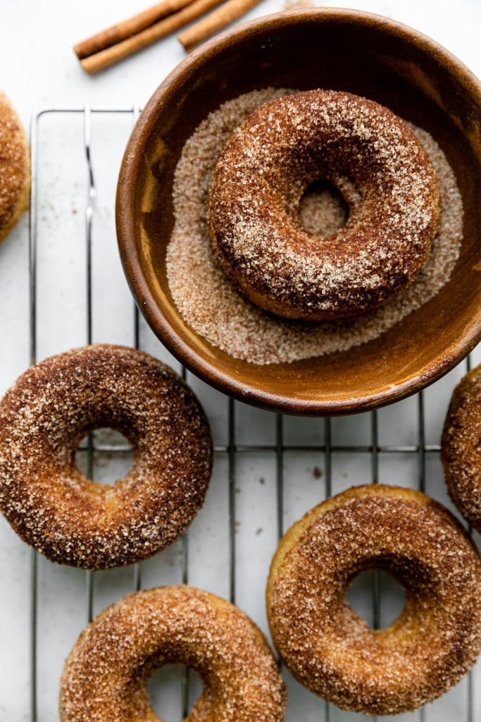 Cinnamon sugar baked donuts on a cooling rack with one donut in a bowl of cinnamon sugar to be coated.
