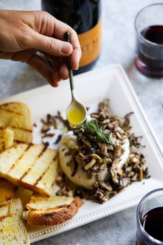 Overhead view of a plate of Burrata Toast with Sautéed Mushrooms and hand drizzling olive oil over the top