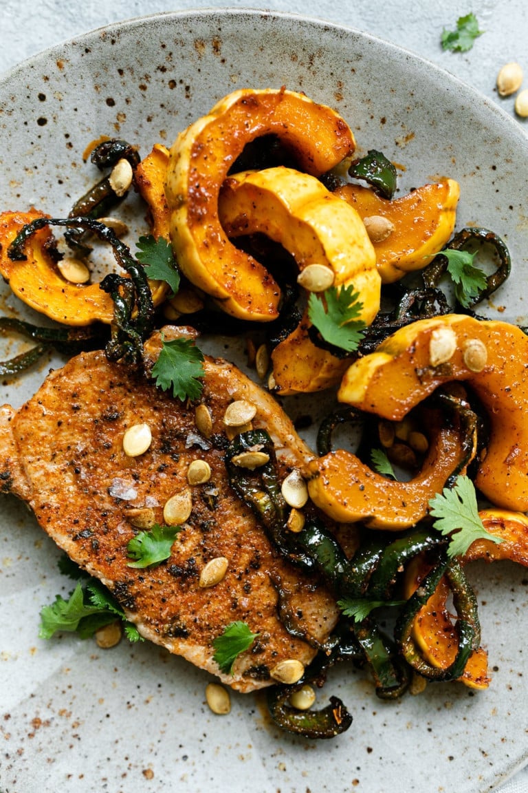Close up view of a plate filled with seasoned pork chops next to roasted delicata squash and topped with pepitas and fresh herbs. 