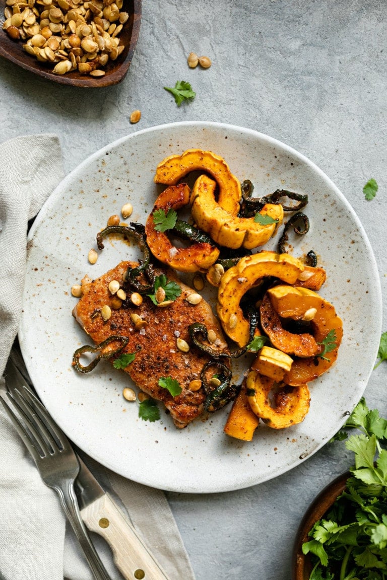 Overhead view of a plate filled with roasted delicata squash, and an oven baked pork chop topped with pumpkin seeds and fresh herbs. 
