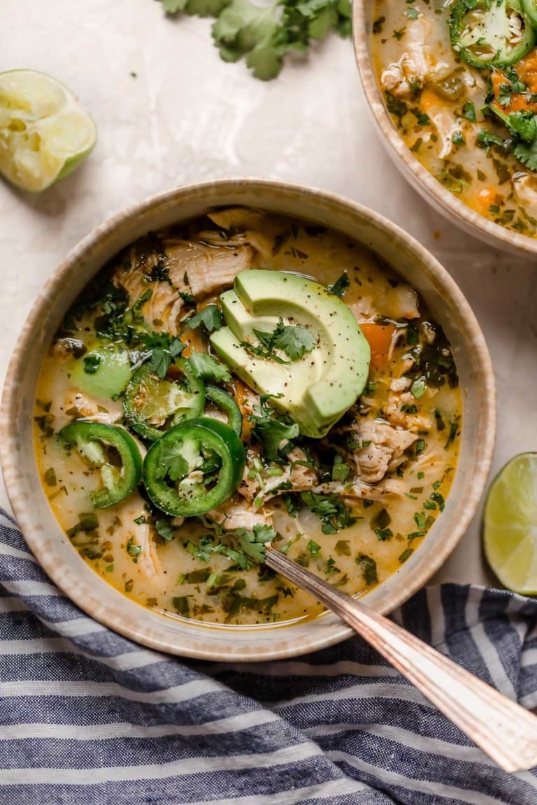 Overhead view of a bowl of white chicken chili topped with fresh jalapenos, sliced avocado, and fresh herbs.
