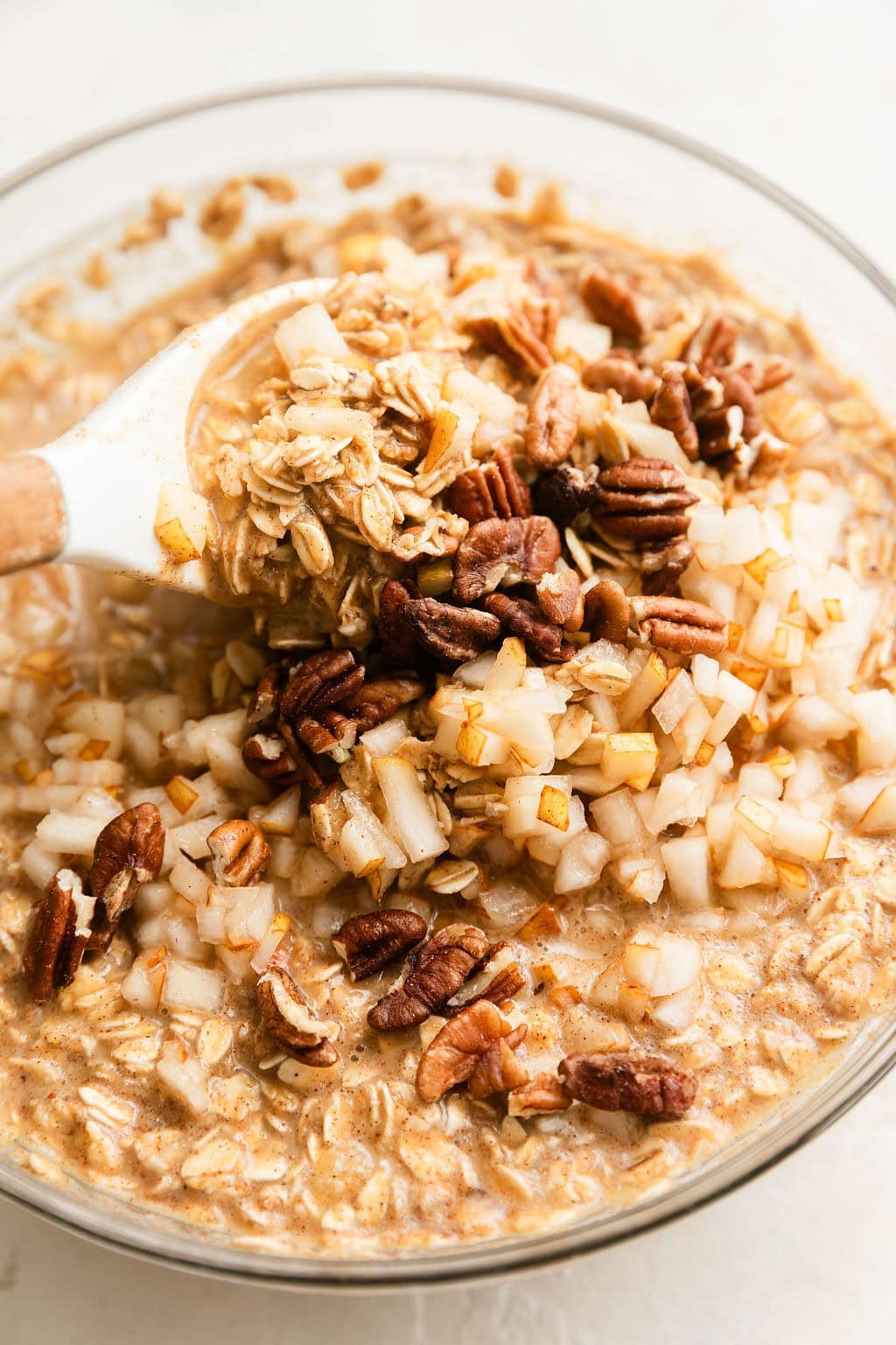 A glass bowl of baked gingerbread oatmeal ingredients with a spoon stirring in pear pieces and pecans.